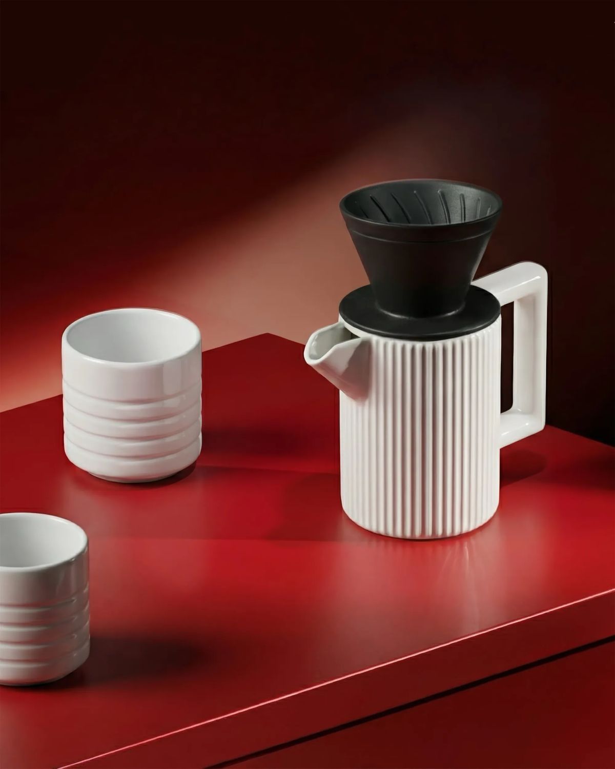 Ribbed white porcelain jug with a black pour-over dripper and two ringed cups on a glossy red table