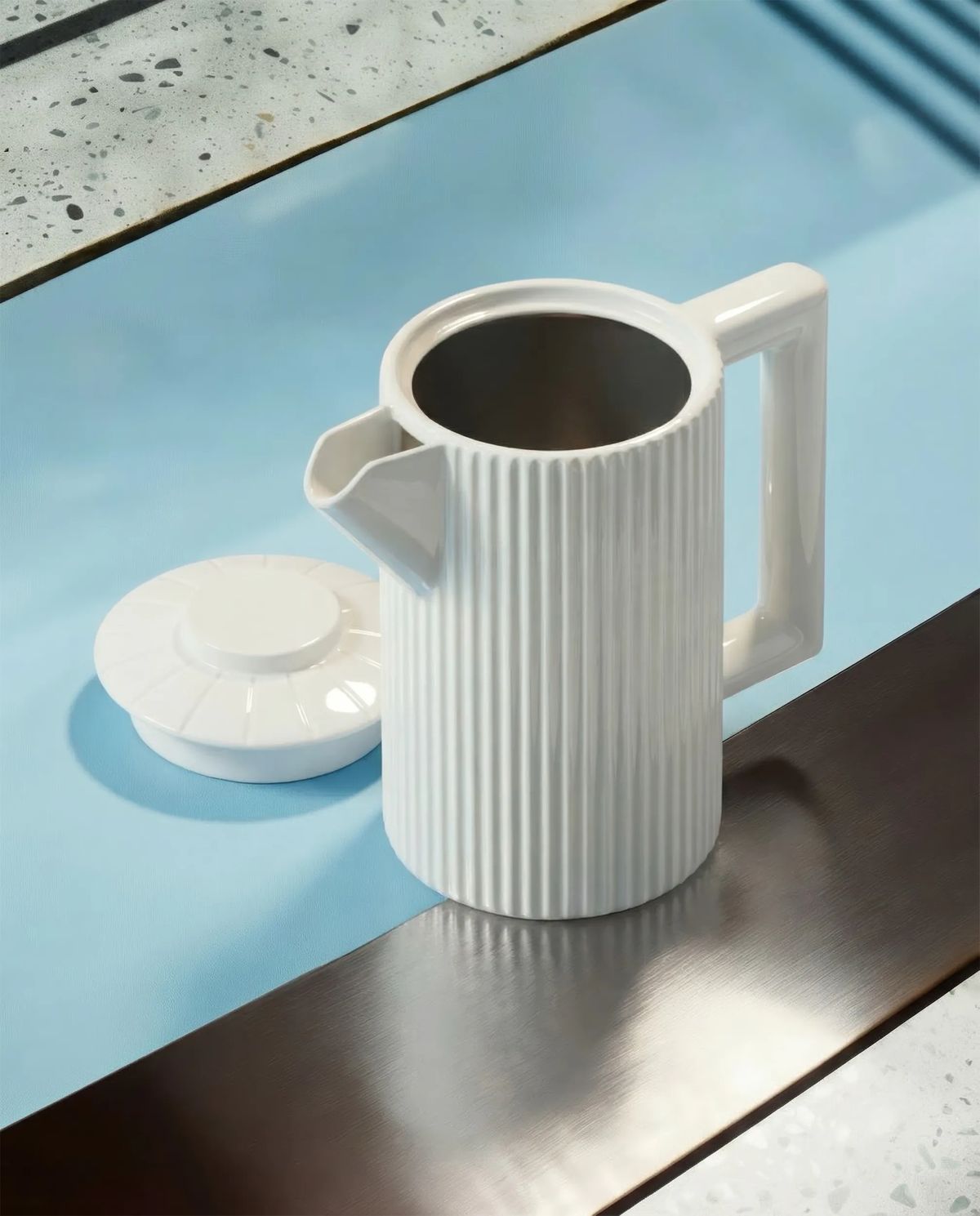 Fluted white porcelain jug with its lid set aside on a blue and brushed steel counter by terrazzo