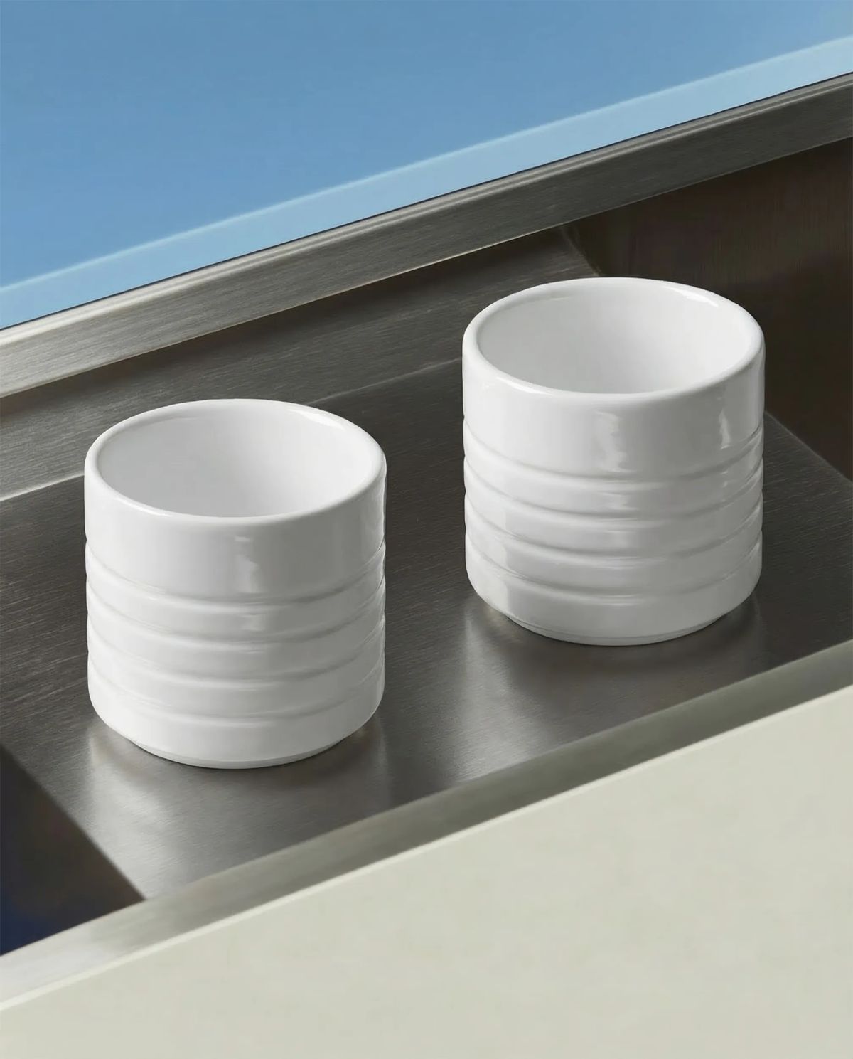 Two ringed white porcelain cups standing in a brushed stainless steel tray against blue