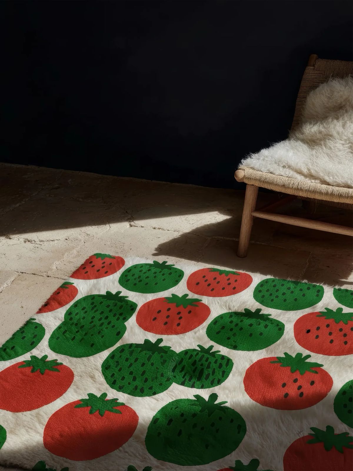 Shaggy cream rug patterned with red and green strawberries on a stone floor beside a sheepskin chair