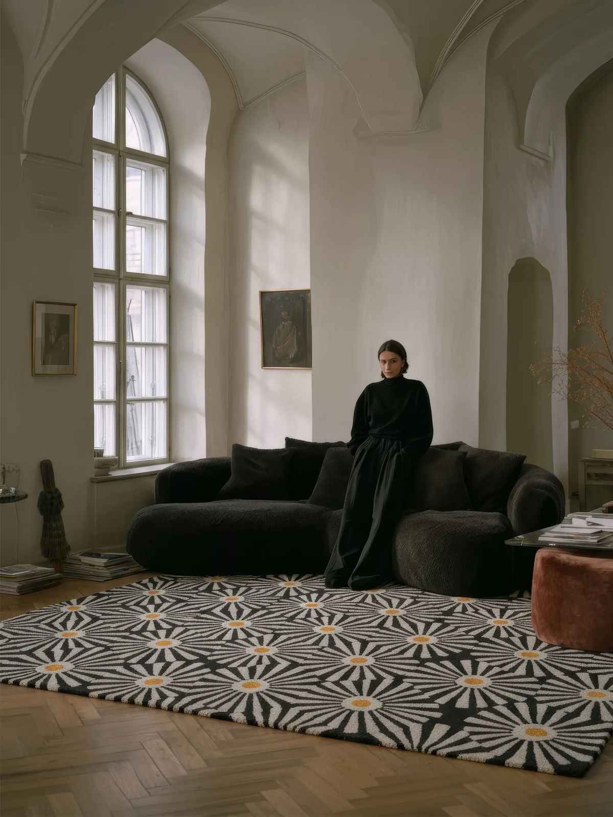 Large black and white daisy-pattern rug before a dark velvet sofa in a vaulted room with arched windows