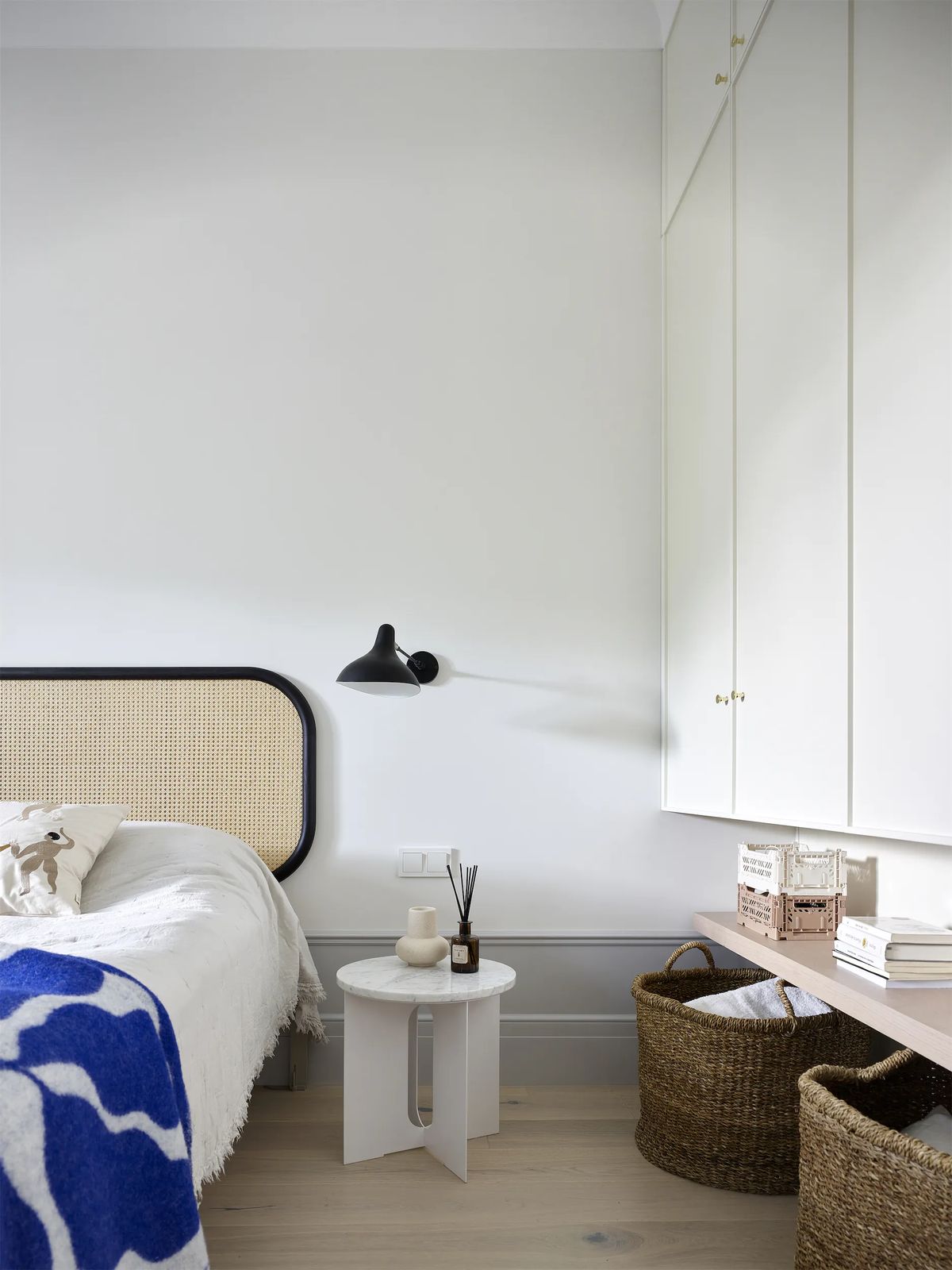 Bedroom corner with cane headboard, blue patterned throw, black wall lamp and white built-in wardrobe
