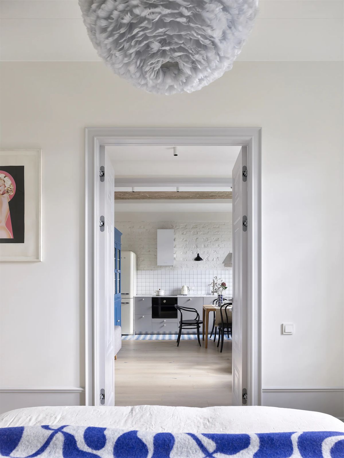 View from the bed through a white doorway to the kitchen, under a grey feather pendant