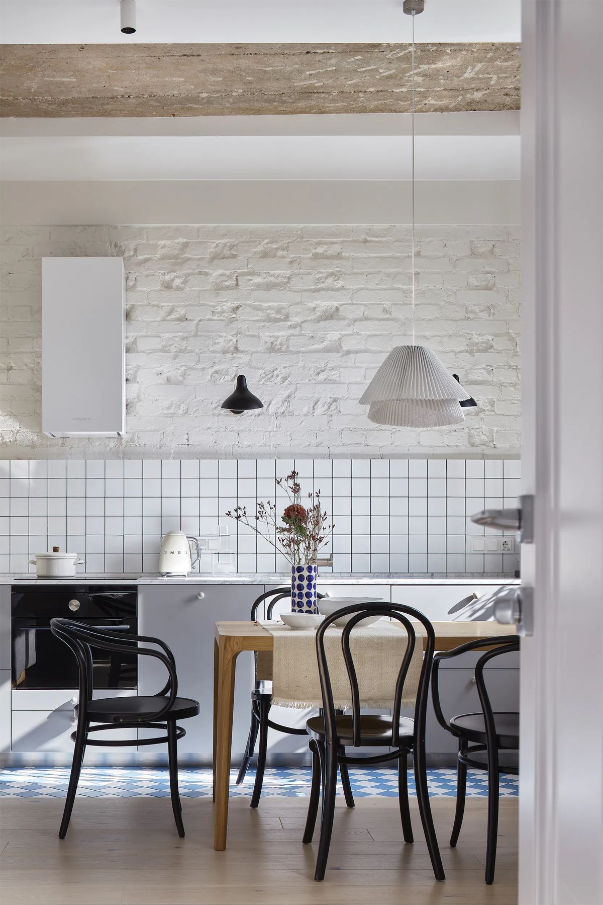 Dining table with black bentwood chairs and pleated pendant before a tiled backsplash and whitewashed brick