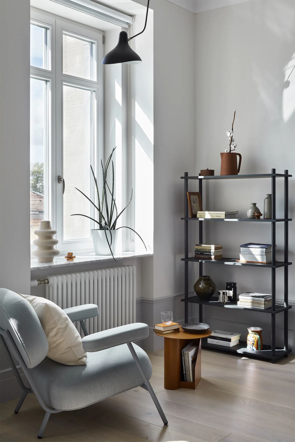 Sunlit corner with pale blue armchair, dark metal shelving, small wooden side table and tall window