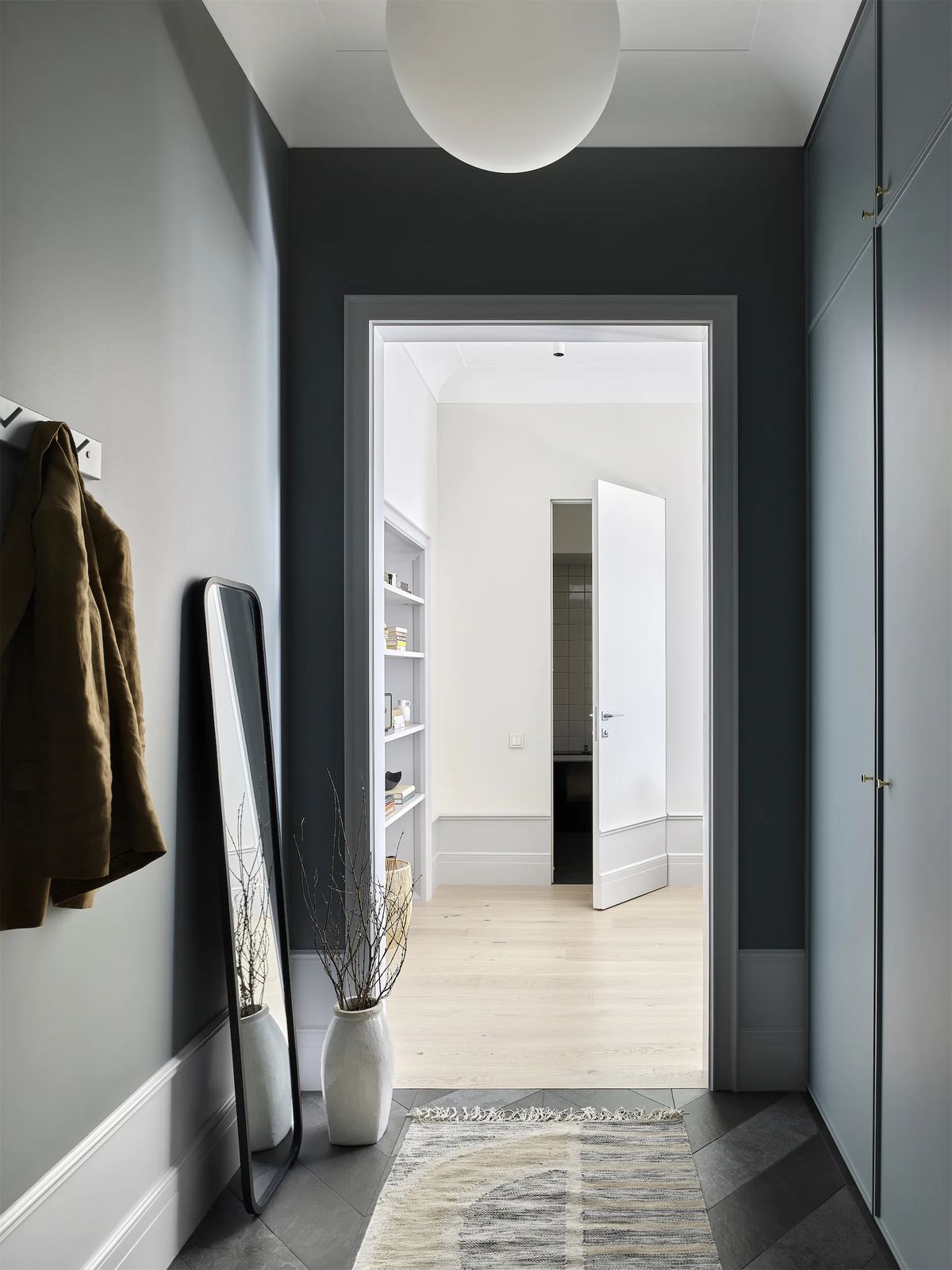 Dark grey hallway with globe pendant, leaning mirror and coat hook, opening onto a bright room