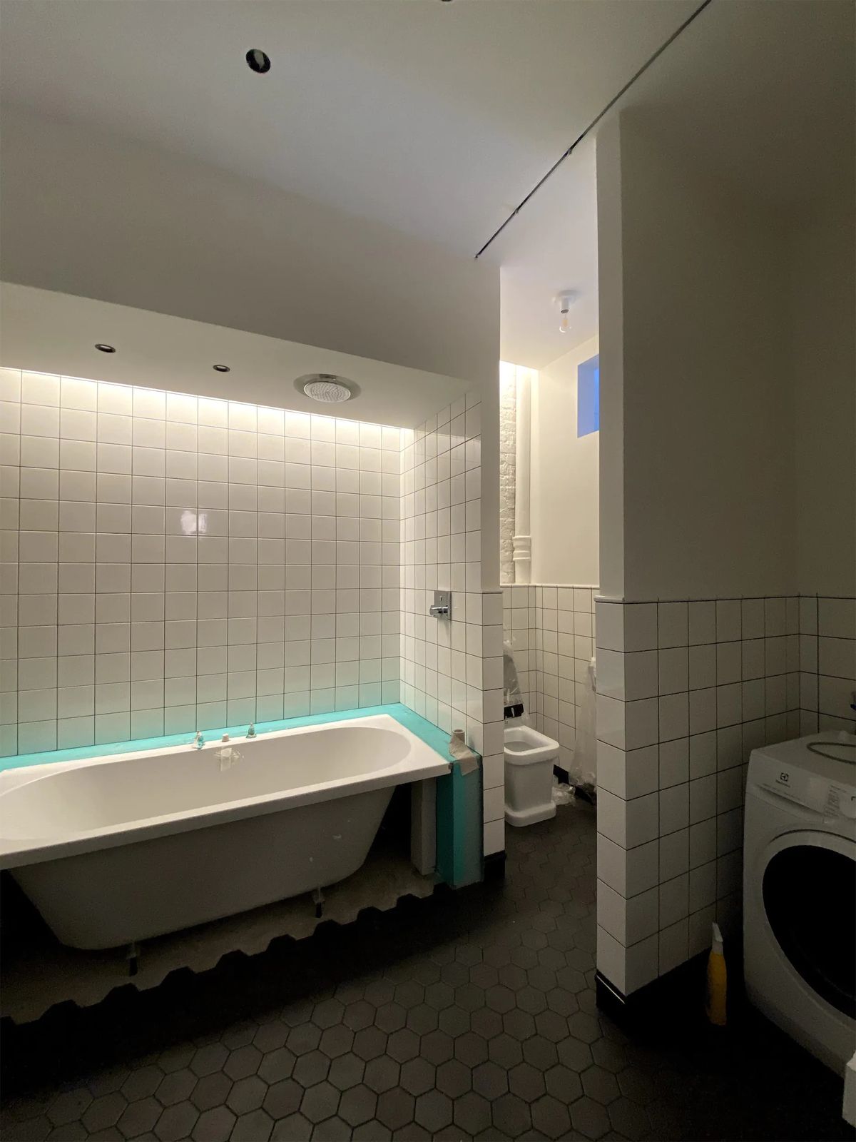 Bathroom during fit-out with a bathtub under backlit white tiles and a washing machine
