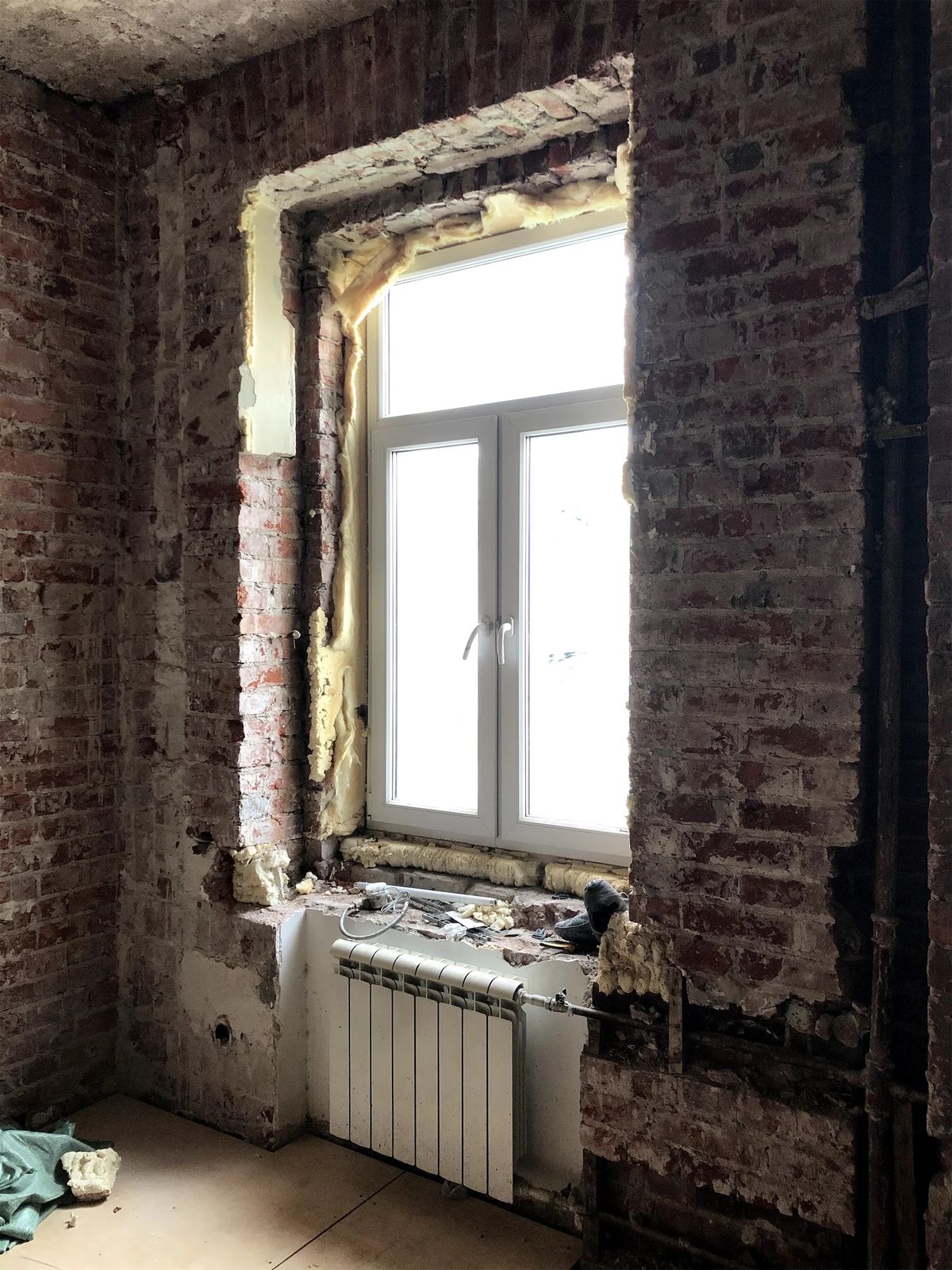 Stripped brick wall around a new white window with foam insulation and a radiator below