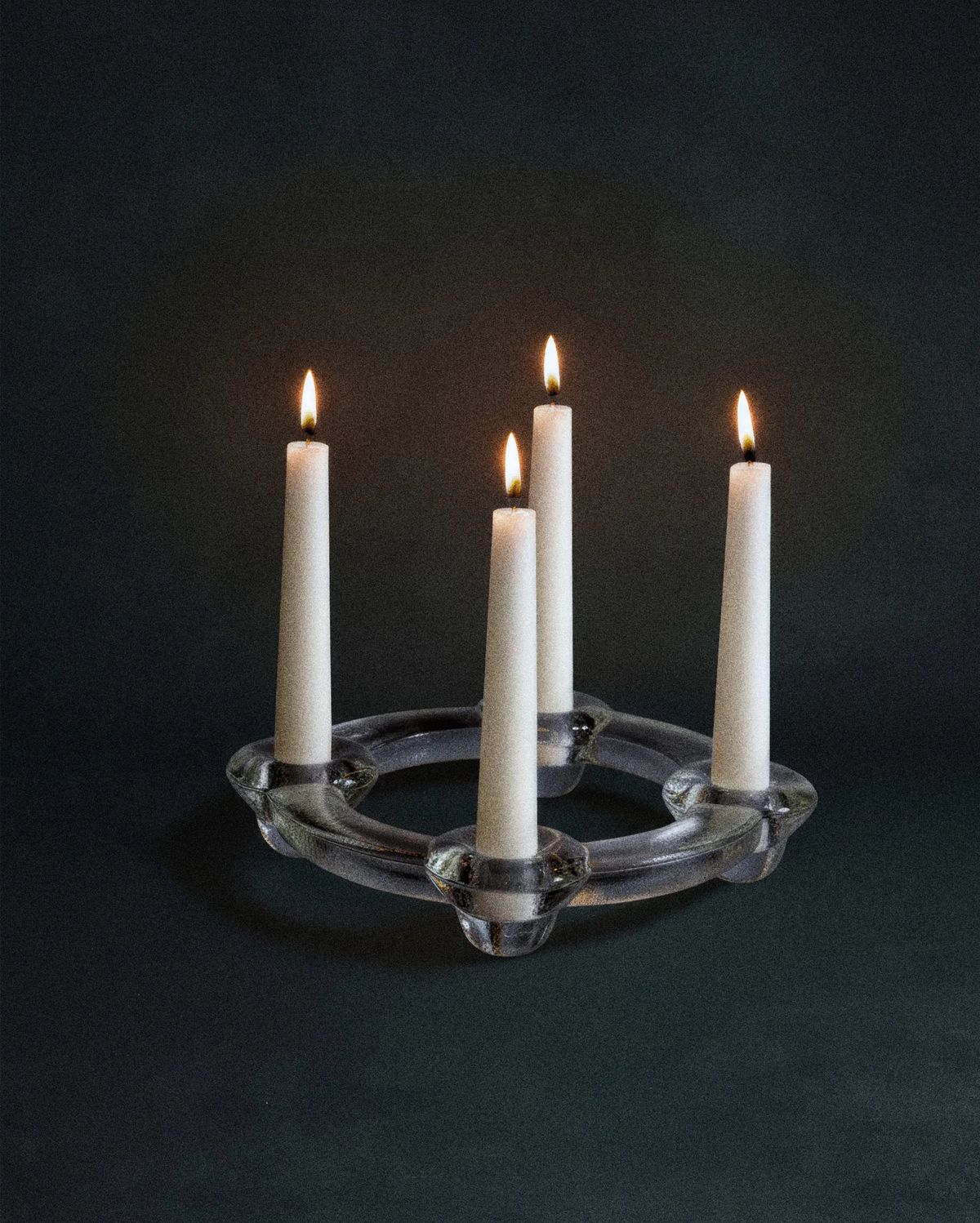 Clear glass ring candleholder holding four lit white taper candles on a dark green backdrop
