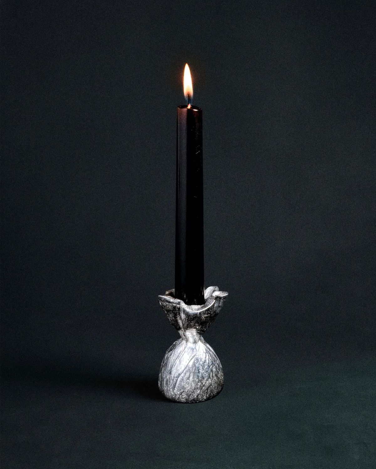 Lit black taper candle in a crumpled silver holder shaped like a tied pouch