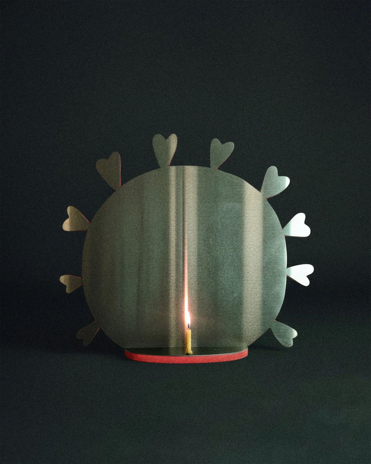 Round brushed-metal shield edged with heart-shaped petals sheltering a tiny lit candle on a red base