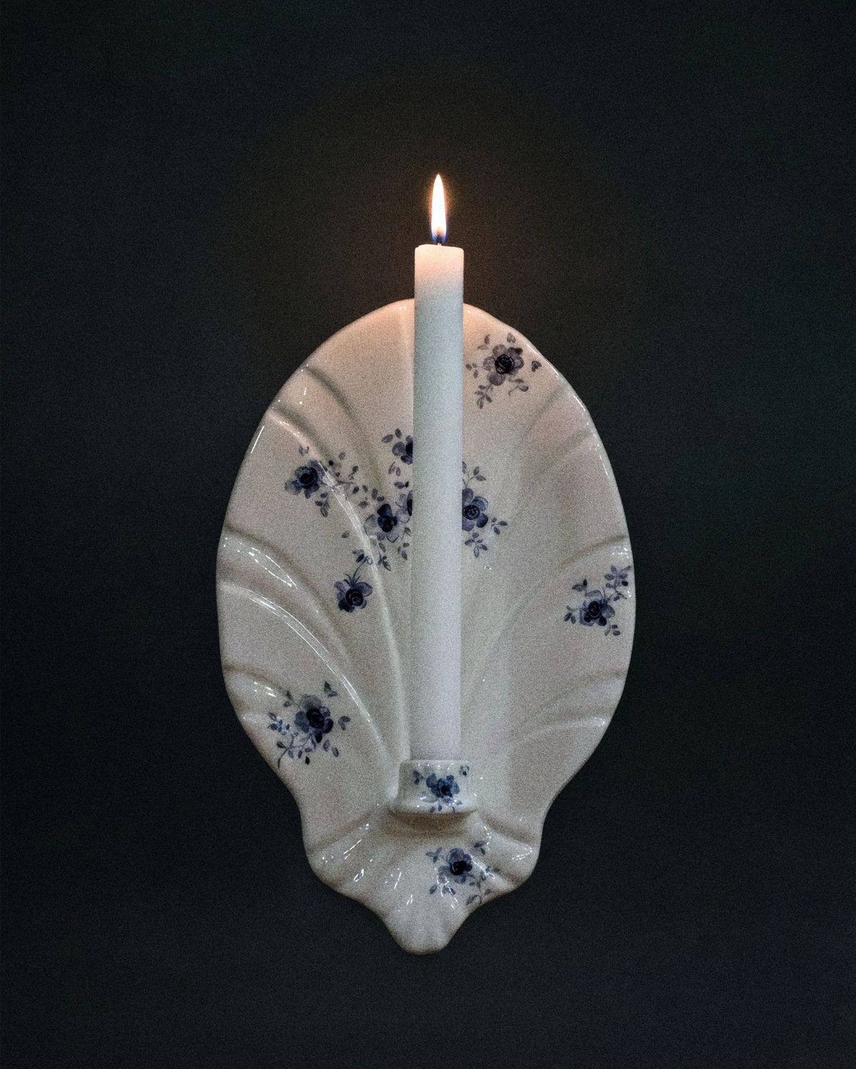 Shell-shaped white ceramic wall sconce with blue floral sprigs holding a single lit candle