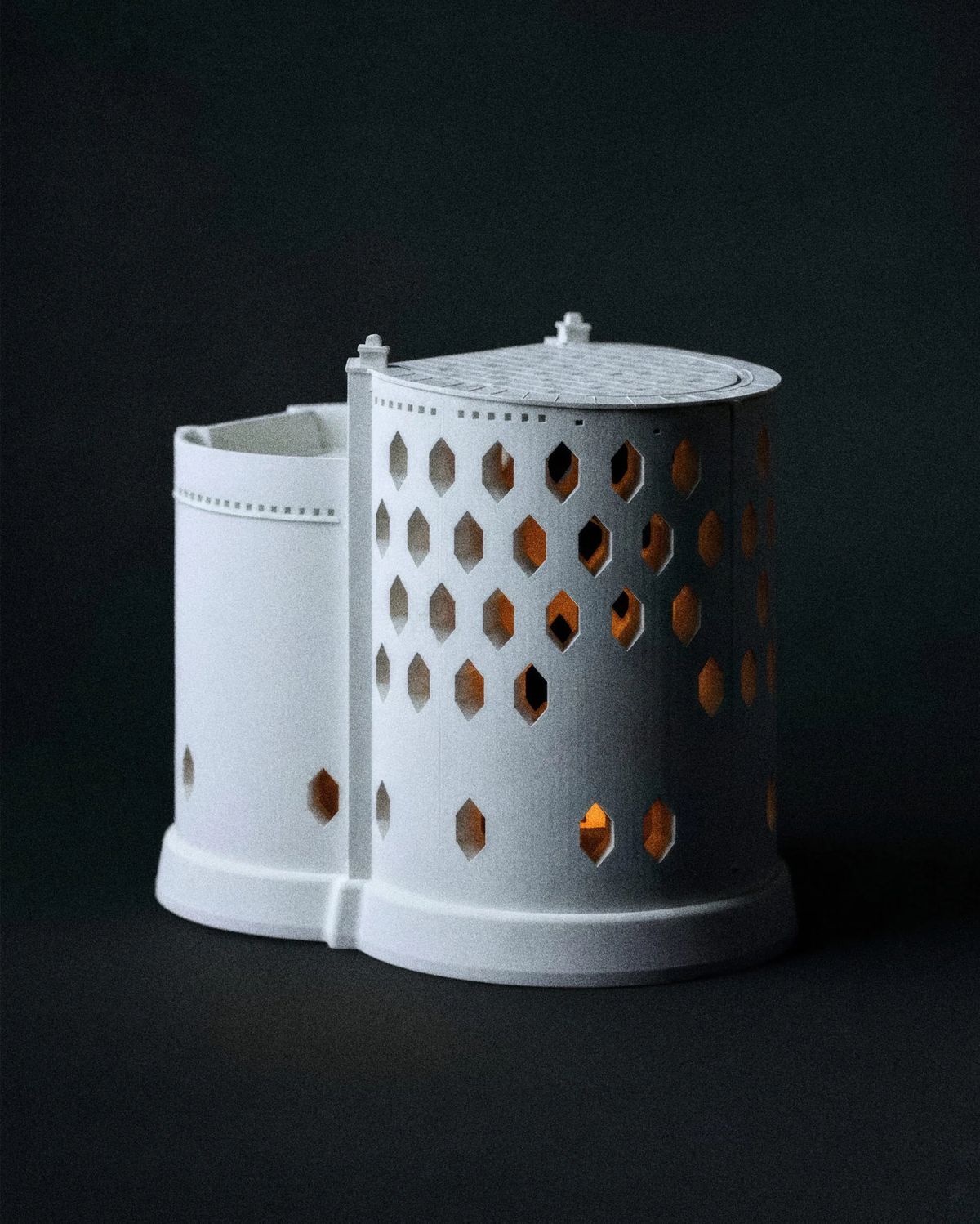 White lantern of two joined cylinders pierced with hexagonal windows glowing amber inside