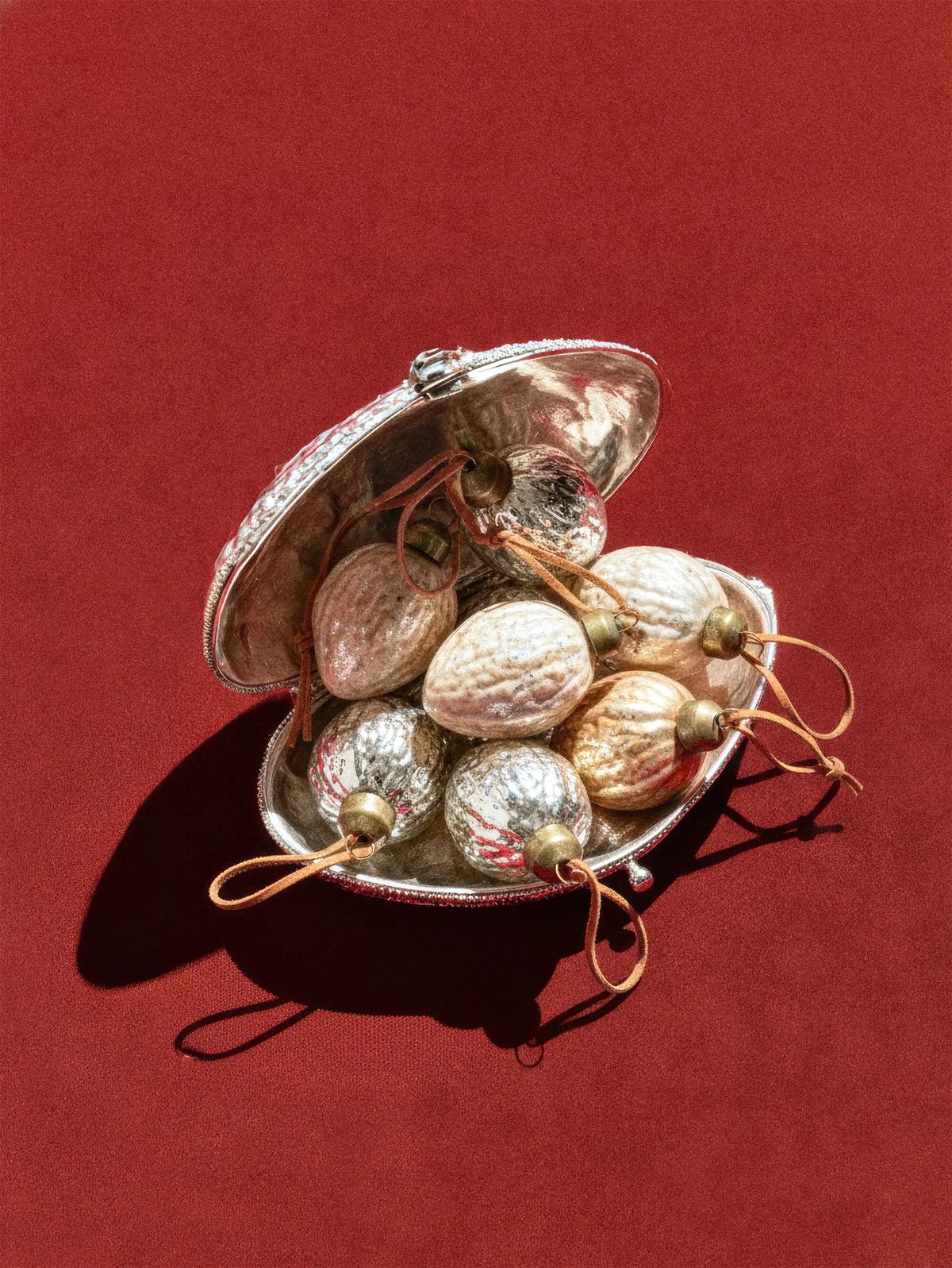 Silver walnut-shaped glass ornaments with leather loops spilling from an open silver shell box on red