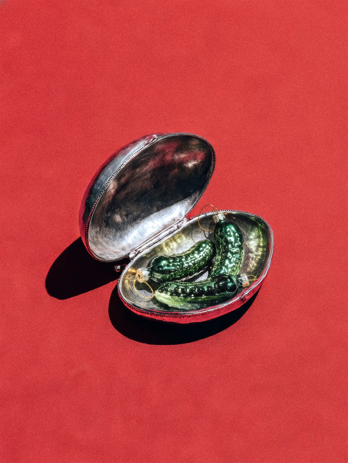 Three green glass pickle ornaments nestled in an open silver shell box on a red background
