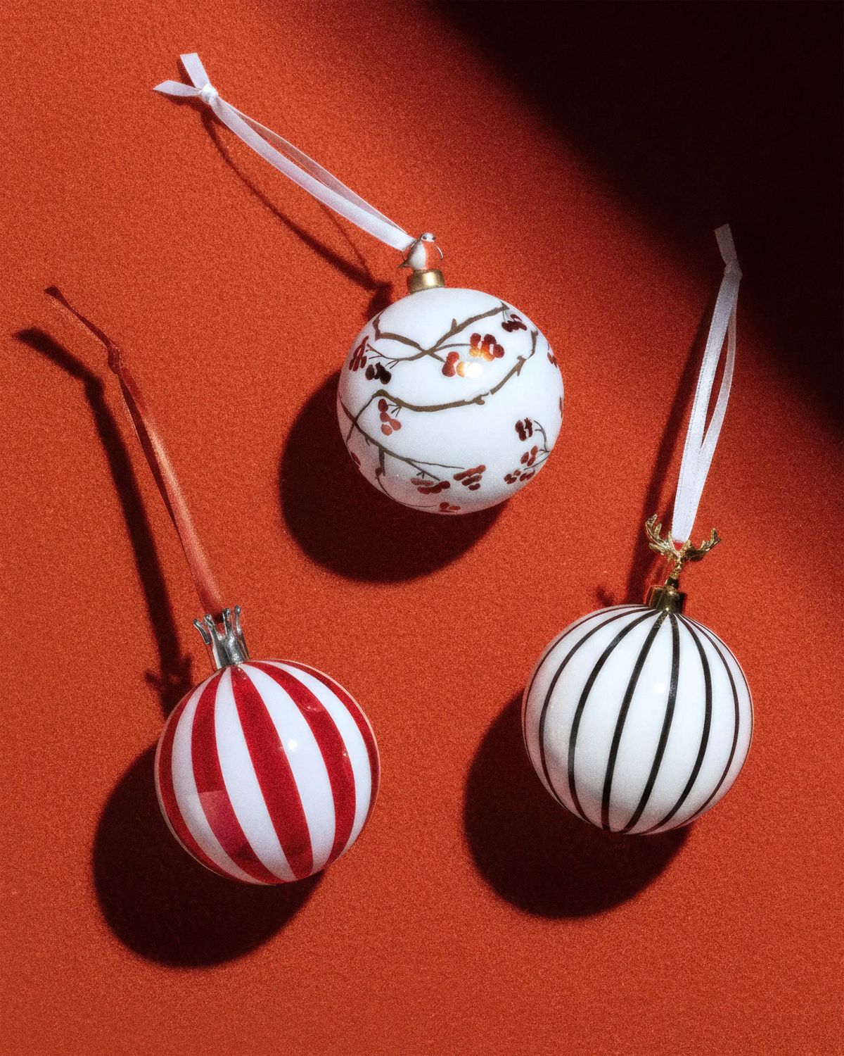 Three white baubles, painted with red stripes, black stripes and berry branches, on orange-red with hard shadows