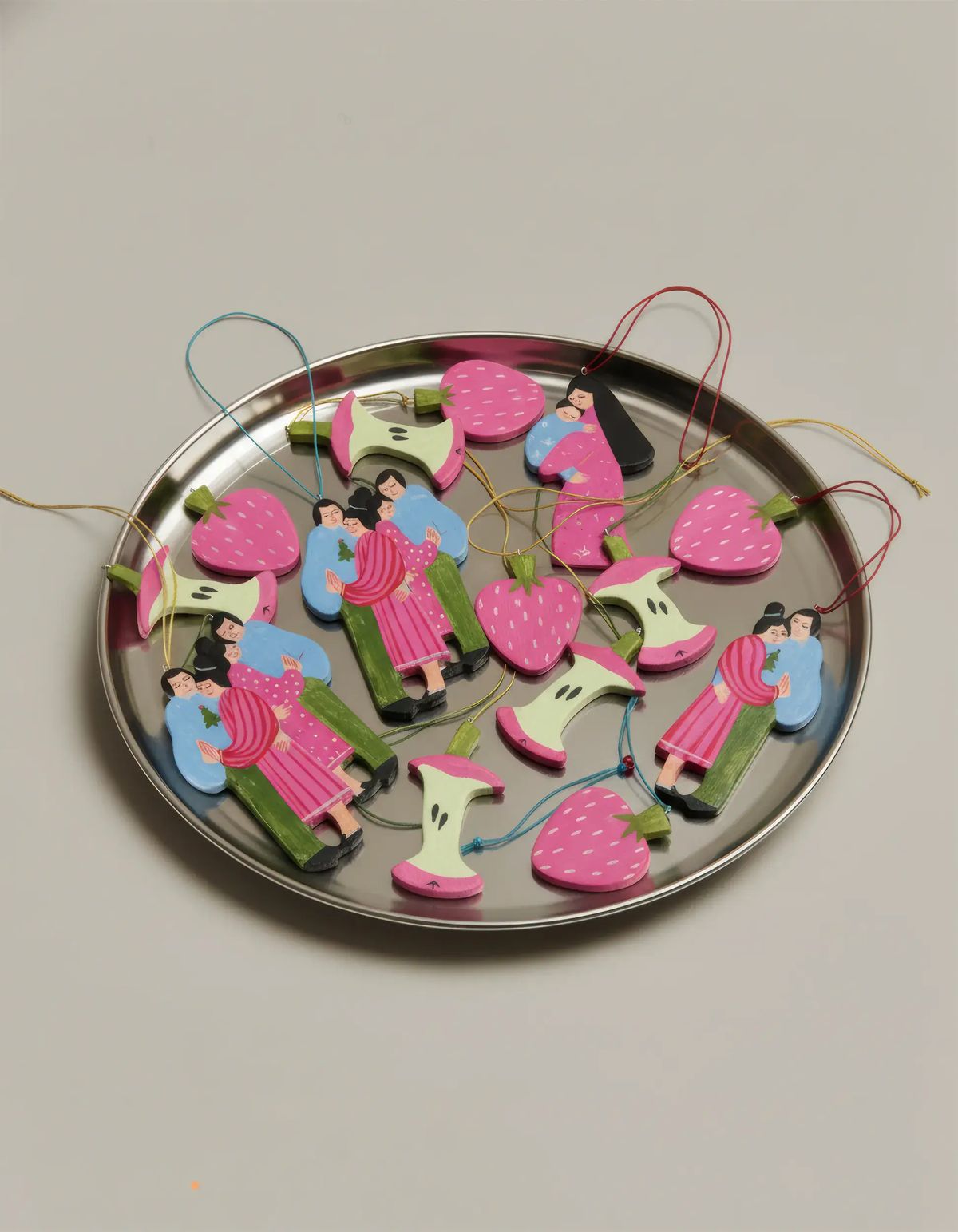 Hand-painted pink strawberry, apple core and embracing couple ornaments with coloured strings on a steel tray