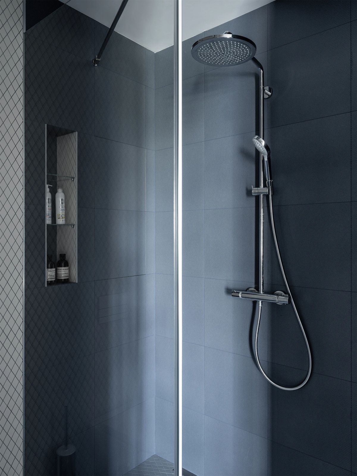 Walk-in shower in dark blue-grey tiles with a chrome rain shower column and a lit wall niche