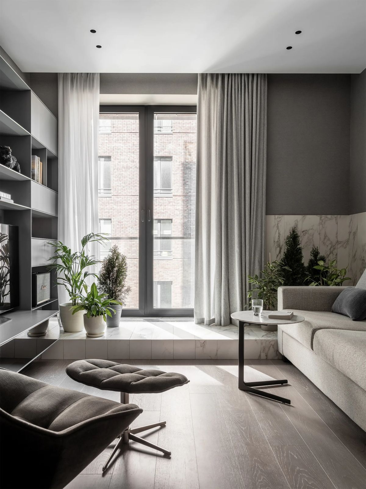 Grey living room with a tall window, linen curtains, potted plants on a marble ledge and lounge chair
