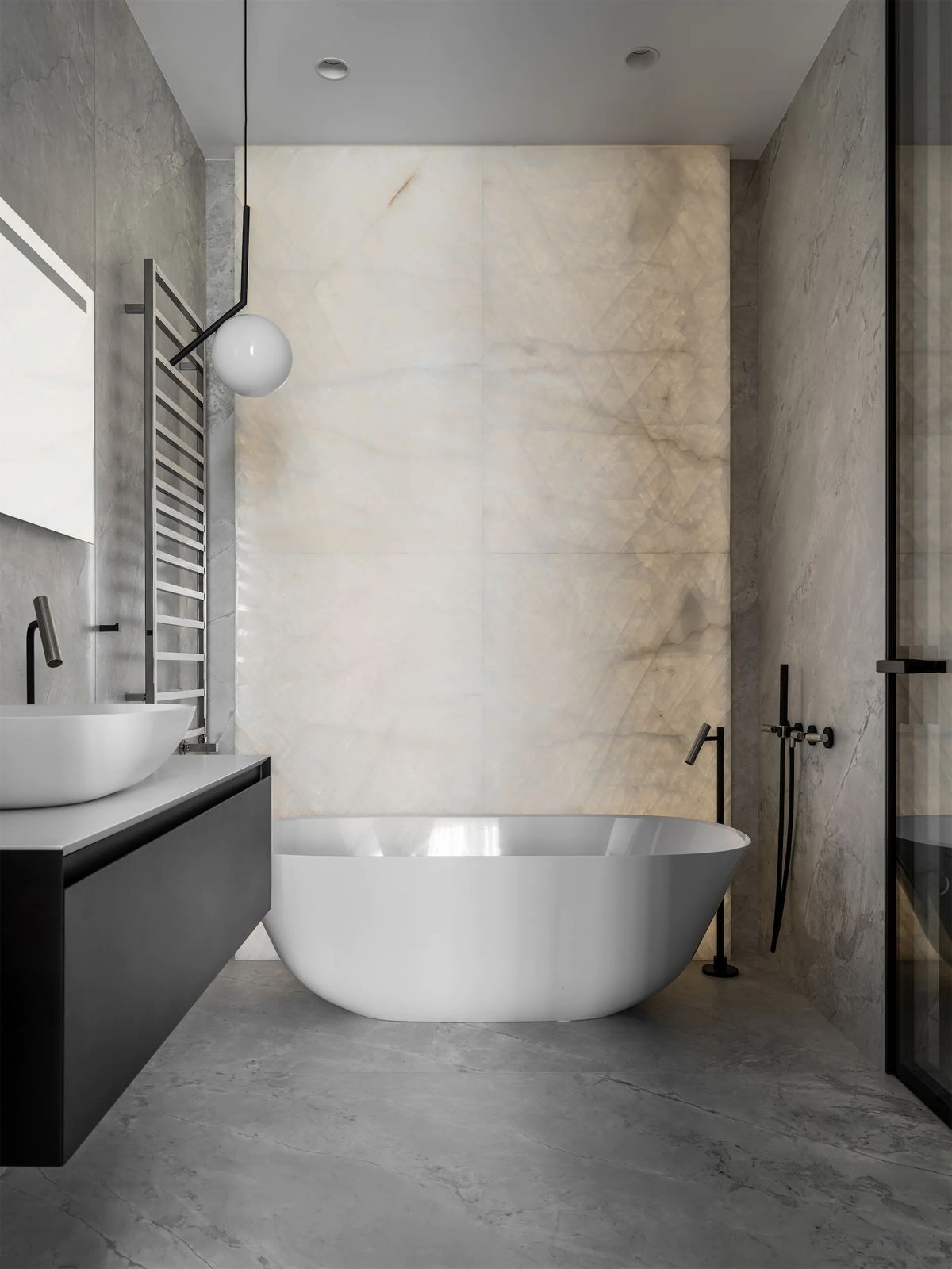 Freestanding white bathtub before a backlit onyx wall, with a globe pendant and grey stone floor