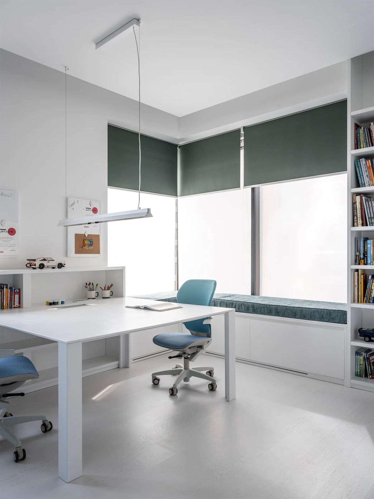 Children's study with a long white desk, blue swivel chairs, green roller blinds and a window seat