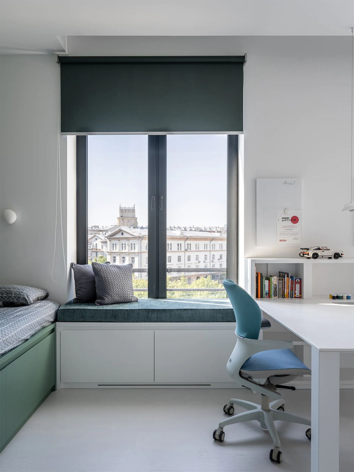 Child's room with a window seat under a green blind, white desk, blue chair and city view