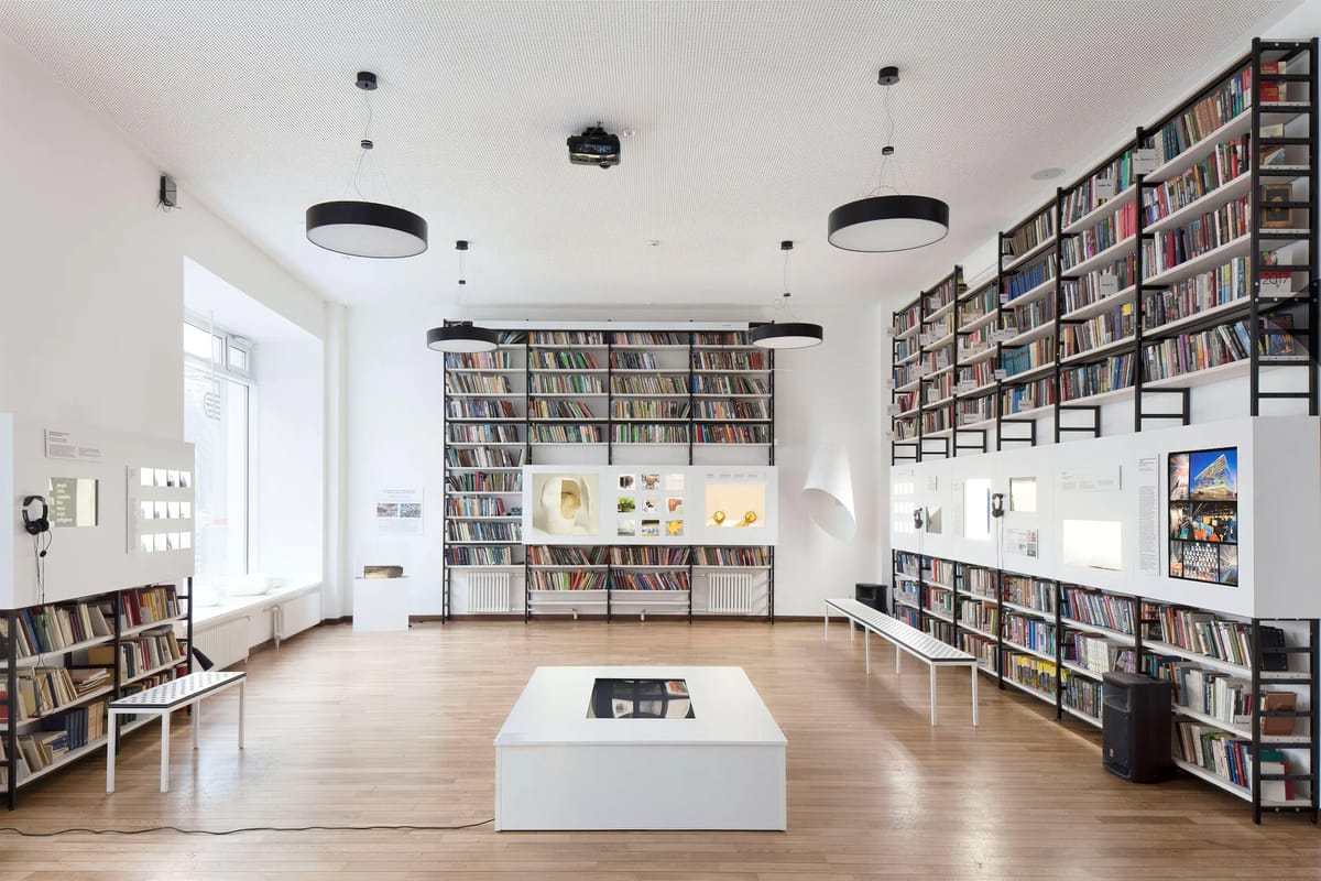 White exhibition room lined with bookshelves, display panels, a low white plinth and benches