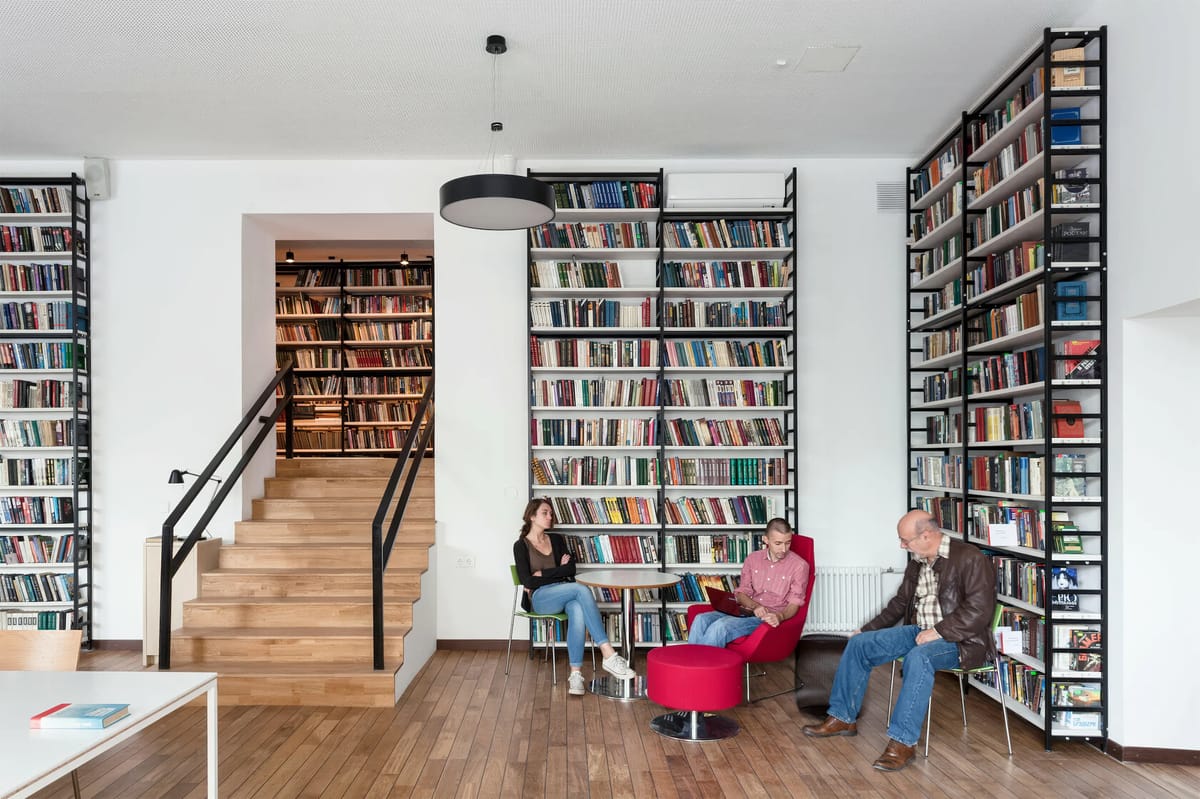 Readers in red chairs between tall bookshelves beside a wide oak staircase leading to more books