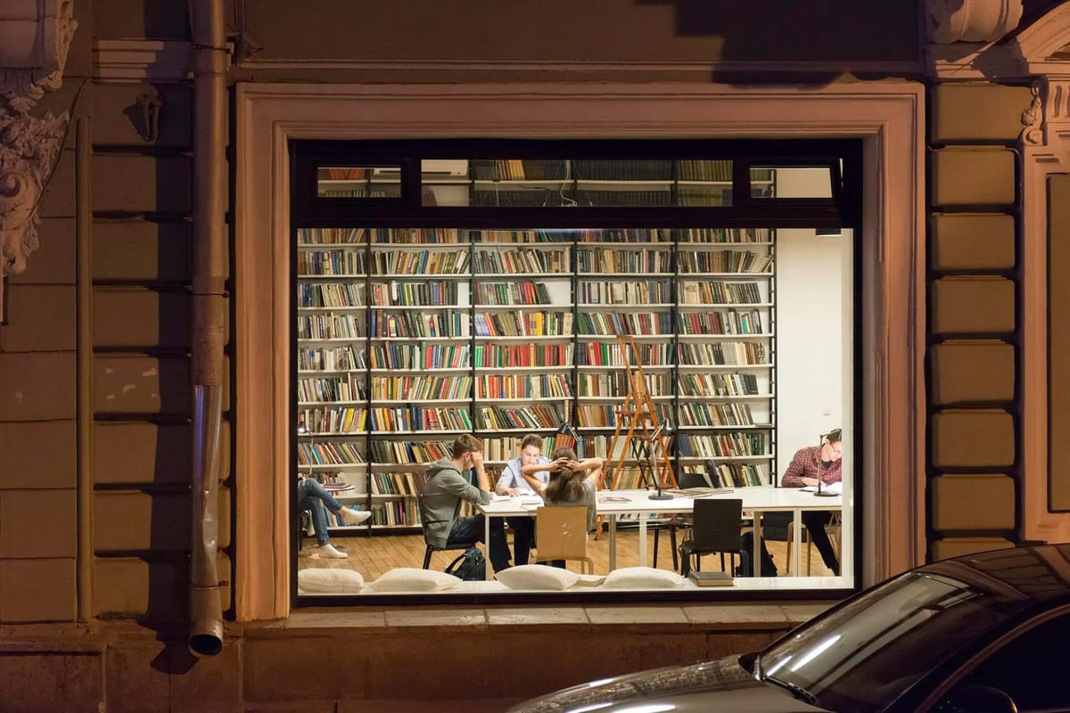 Evening view through a large window into a lit reading room with bookshelves and a long shared table