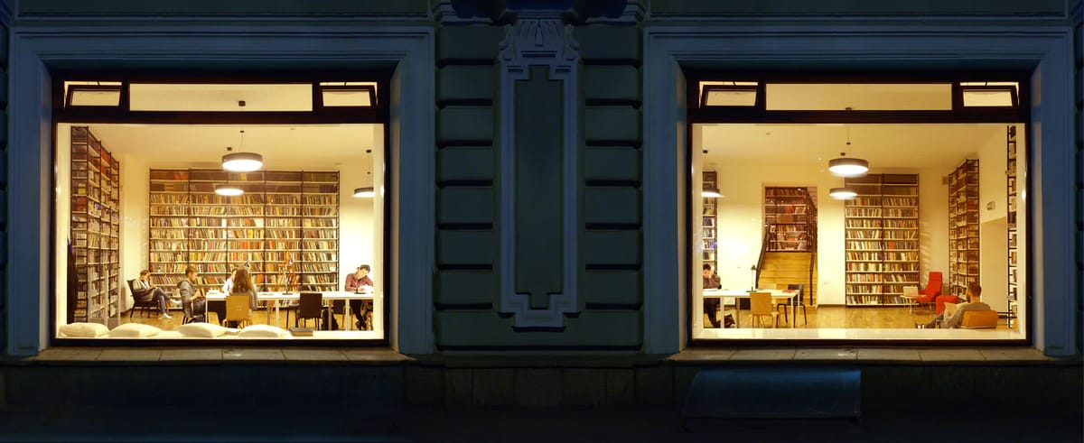 Two lit library windows at night in a historic facade, showing bookshelves, tables and readers