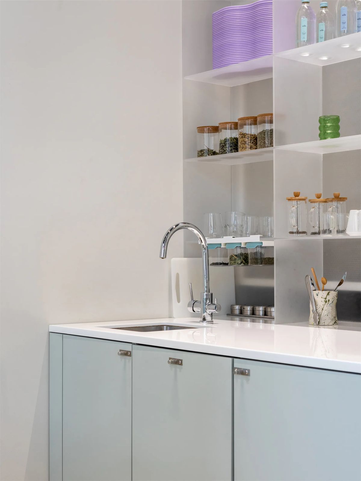 Staff kitchenette with sage cabinets, chrome tap and open metal shelves holding glass jars