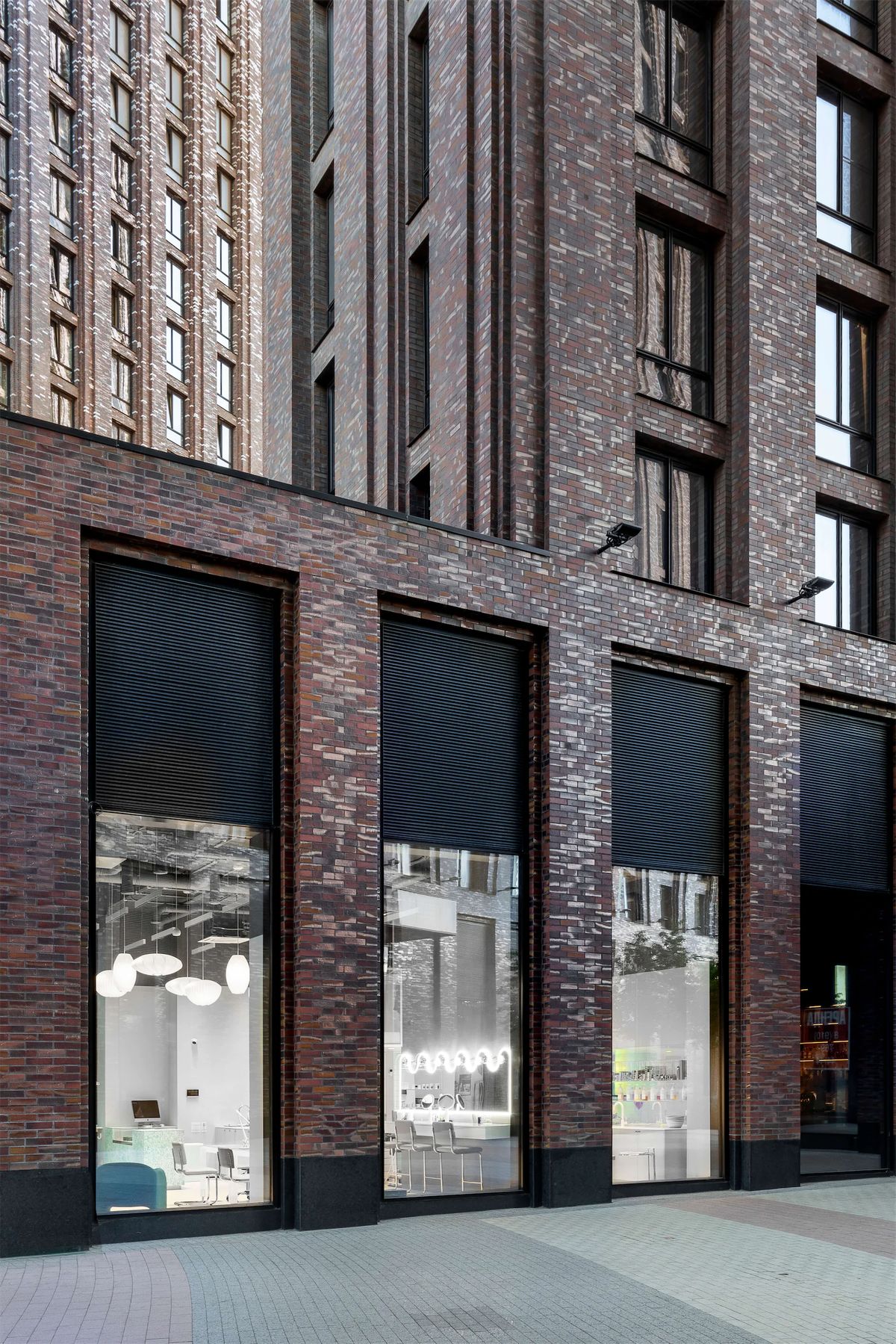 Dark brick residential tower with tall shopfront windows glowing with the white salon interior