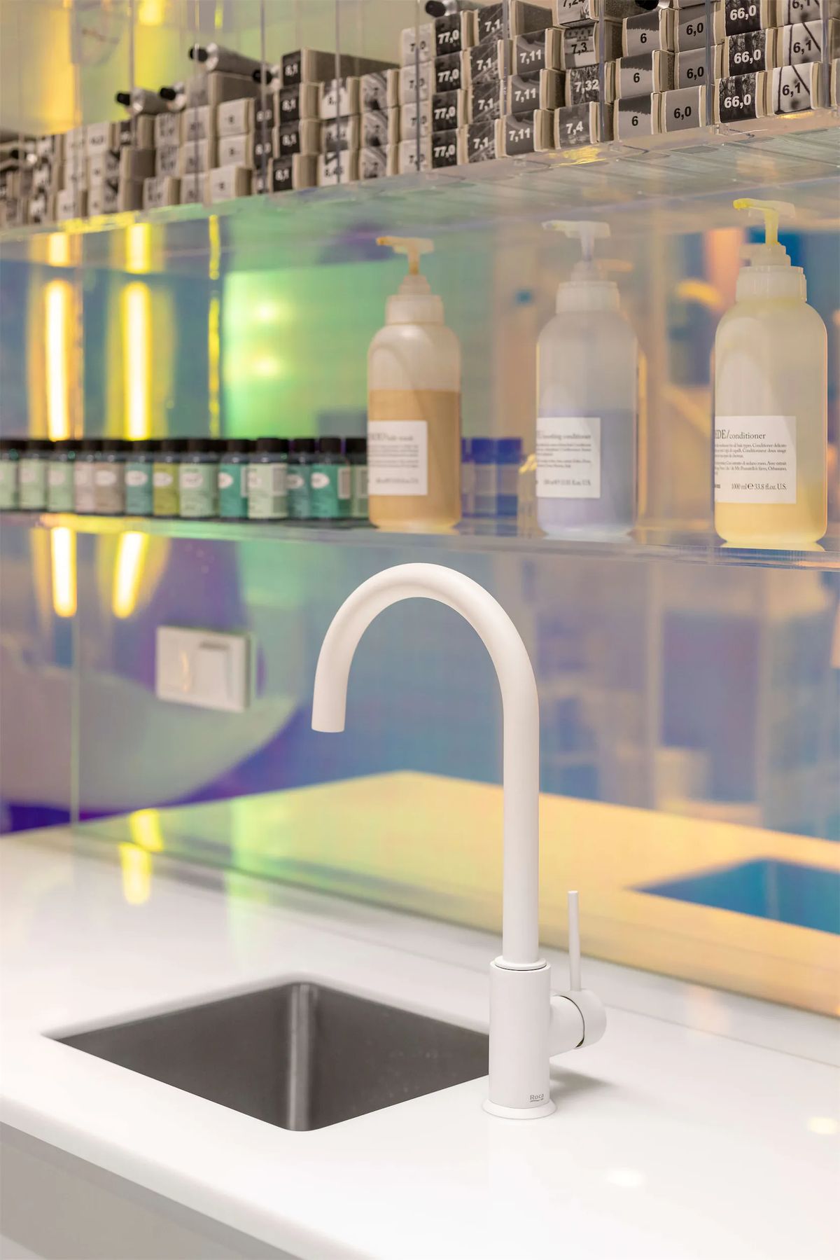 White gooseneck tap over a steel sink below iridescent glass shelves of dyes and conditioner bottles