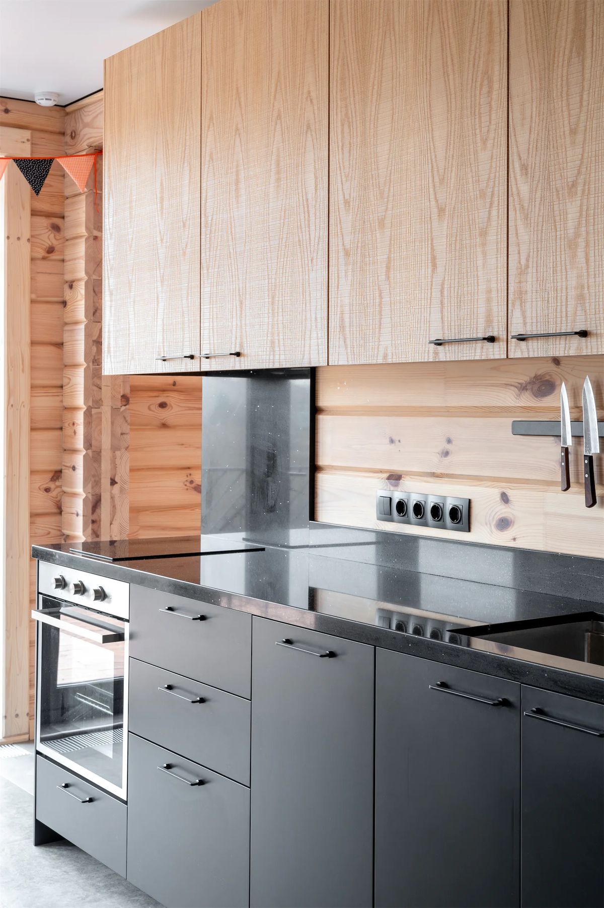 Kitchen with ash-veneer wall cabinets, black worktop and graphite base units against log walls