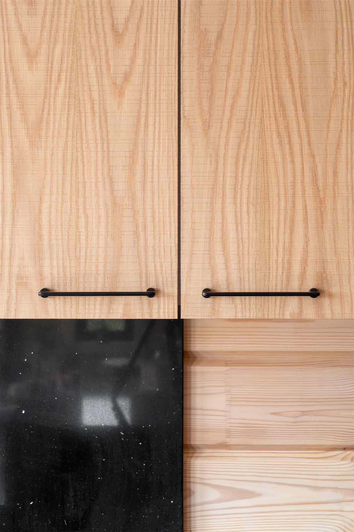 Detail of wood-veneer cabinet doors with black bar handles above black stone and pine boards