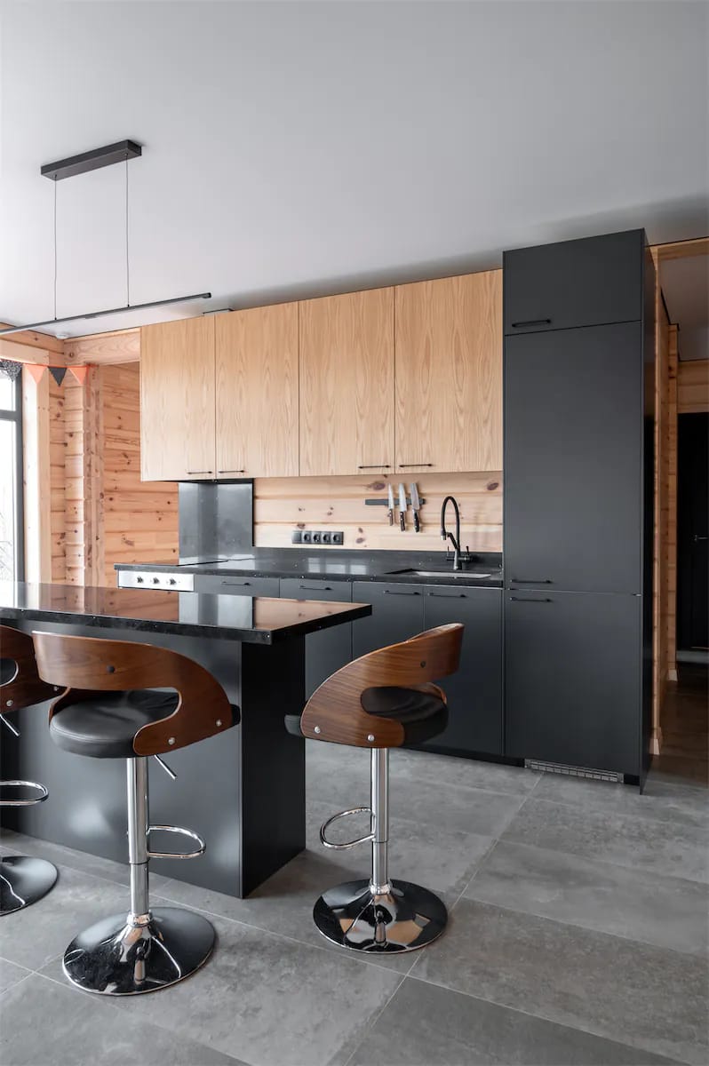 Open kitchen in a log house with a black island, wooden bar stools and grey stone floor tiles