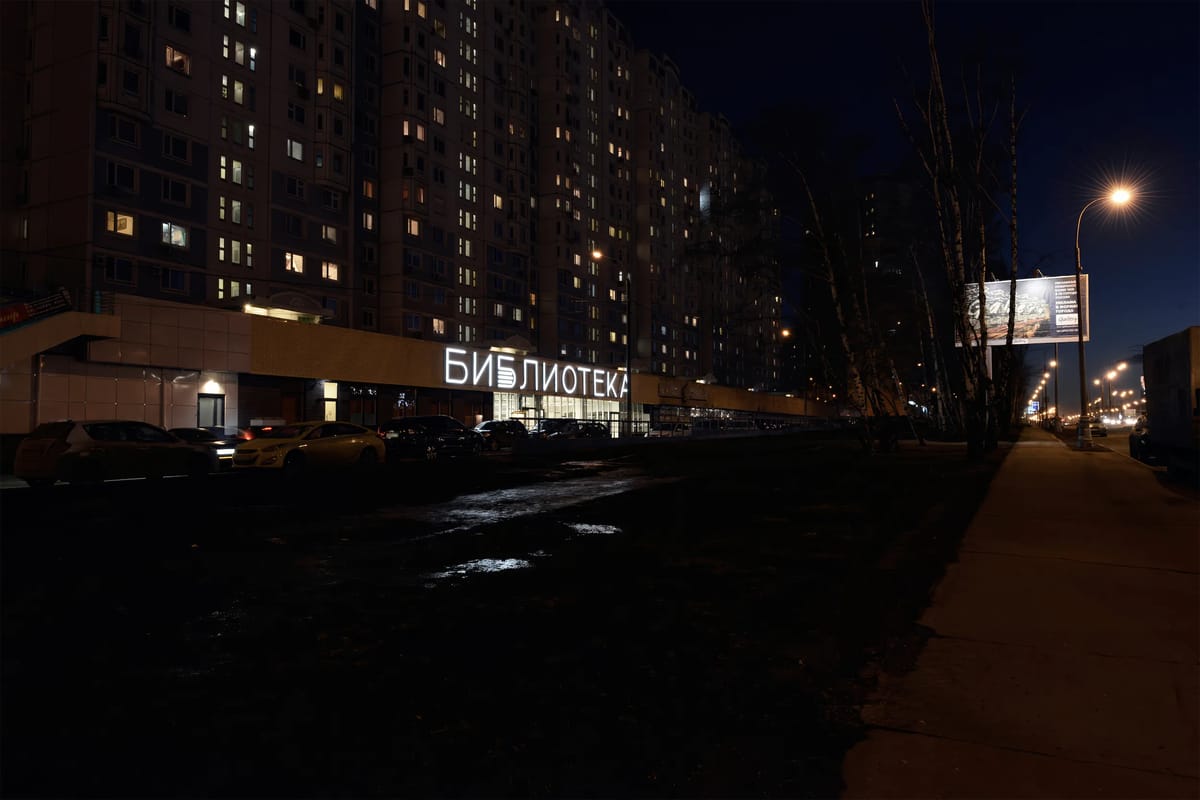 Night view along a wide avenue to a glowing library sign below a high-rise apartment block