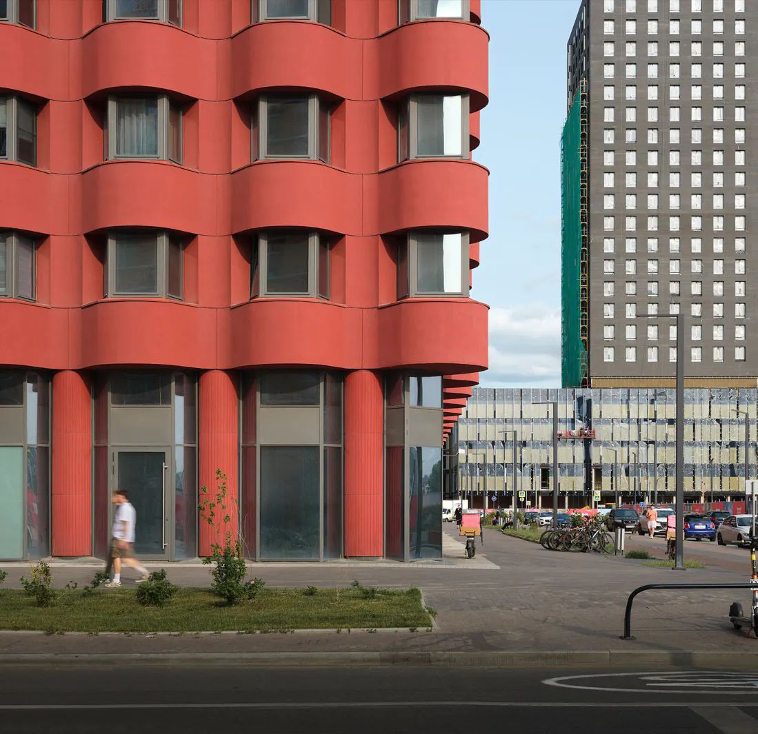 Red residential facade with rounded bay windows and columns at street level beside a grey tower