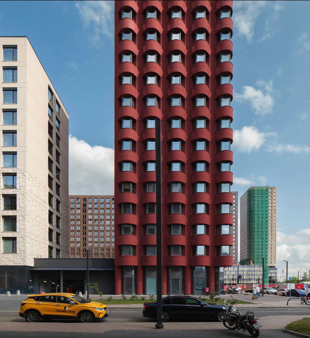 Slim red tower with stacked curved bay windows rising between neighbouring blocks under a blue sky
