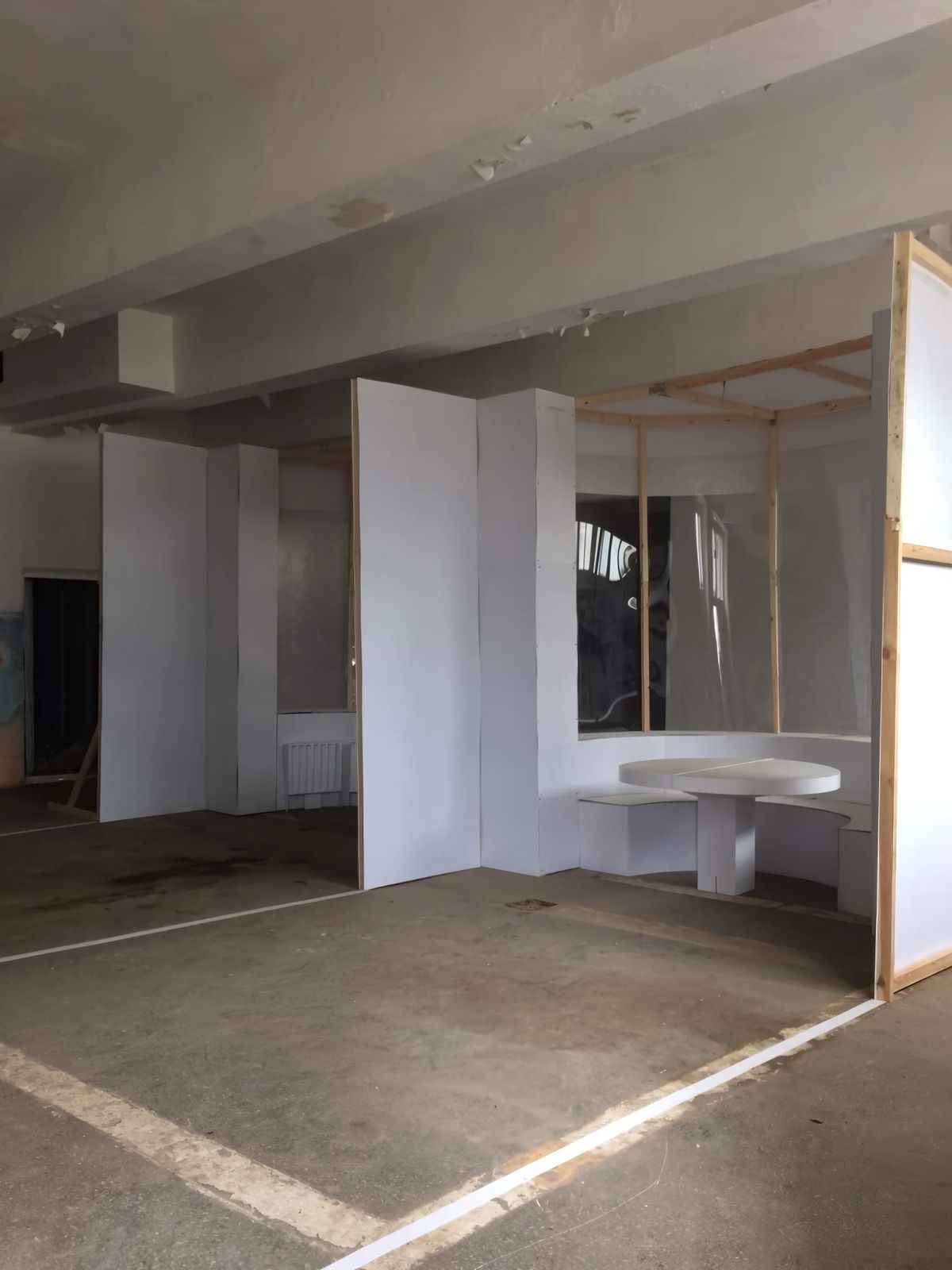 Full-scale mock-up of a curved bay with built-in bench and round table made of white boards and timber